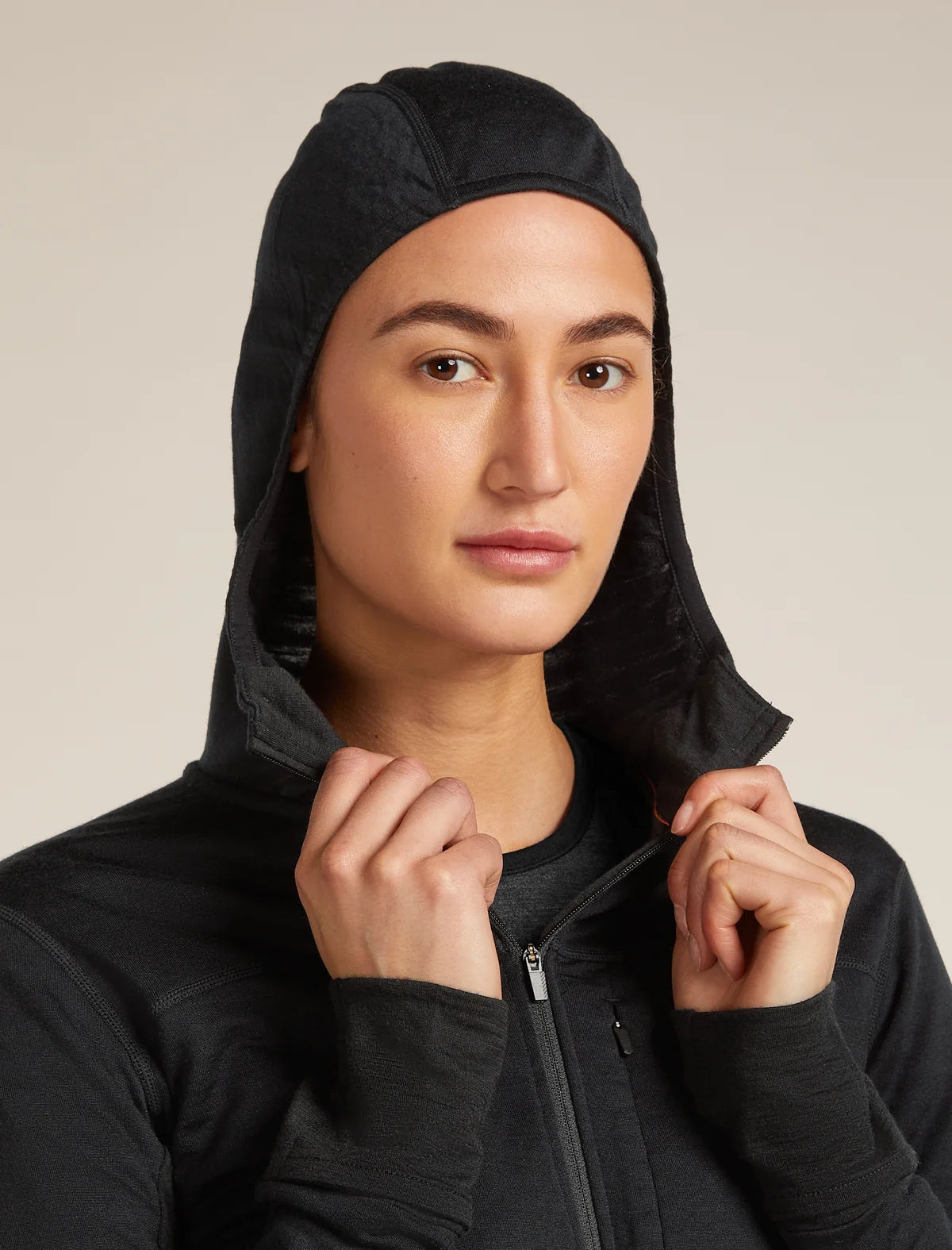 A close-up of a model wearing a black hooded zip-up jacket, holding the hood up. The background is a solid, warm off-white studio backdrop. - Icebreaker Merino Blend 300 RealFleece Descender Long Sleeve Zip Hoodie (Women's) - Black