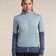 A woman modeling a blue full-zip hooded athletic jacket over a dark gray shirt, paired with gray pants, against a plain light beige studio background. - Icebreaker Merino Blend 300 RealFleece Descender Long Sleeve Zip Hoodie (Women's) - blue, light blue, dawn/zinc blue