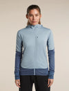 A woman modeling a blue full-zip hooded athletic jacket over a dark gray shirt, paired with gray pants, against a plain light beige studio background. - Icebreaker Merino Blend 300 RealFleece Descender Long Sleeve Zip Hoodie (Women's) - blue, light blue, dawn/zinc blue