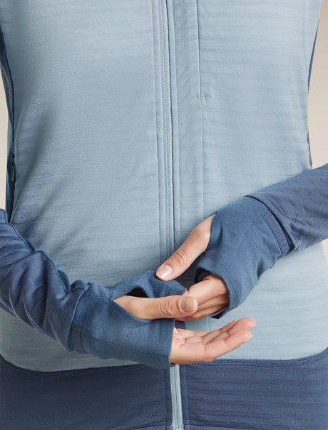 Close-up of a person wearing a blue long-sleeve athletic zip jacket, showing the cuffs and torso against a plain light beige studio background. - Icebreaker Merino Blend 300 RealFleece Descender Long Sleeve Zip Hoodie (Women's) - blue,  light blue, dawn/ zinc blue