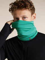 A close-up of a man wearing a bright green neck gaiter pulled up over his lower face, paired with a black zip-up jacket against a plain beige background. - Icebreaker Merino 200 Flexi Chute (Unisex) - Fresh
