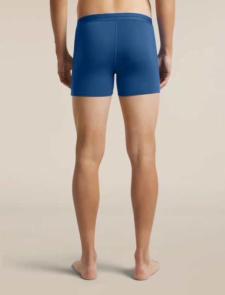 A man viewed from the back wearing blue boxer briefs, standing barefoot against a plain beige studio background. - Icebreaker Merino 150 Anatomica Boxers (Men's) - Lazurite