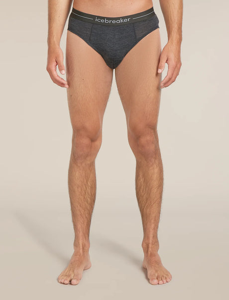 A man wearing dark gray heathered briefs with a black waistband featuring the white 'icebreaker' logo, shown from the waist down against a light beige studio background. - Icebreaker Merino 150 Anatomica Briefs (Men's) - Jet Heather