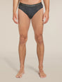 A man wearing dark gray heathered briefs with a black waistband featuring the white 'icebreaker' logo, shown from the waist down against a light beige studio background. - Icebreaker Merino 150 Anatomica Briefs (Men's) - Jet Heather