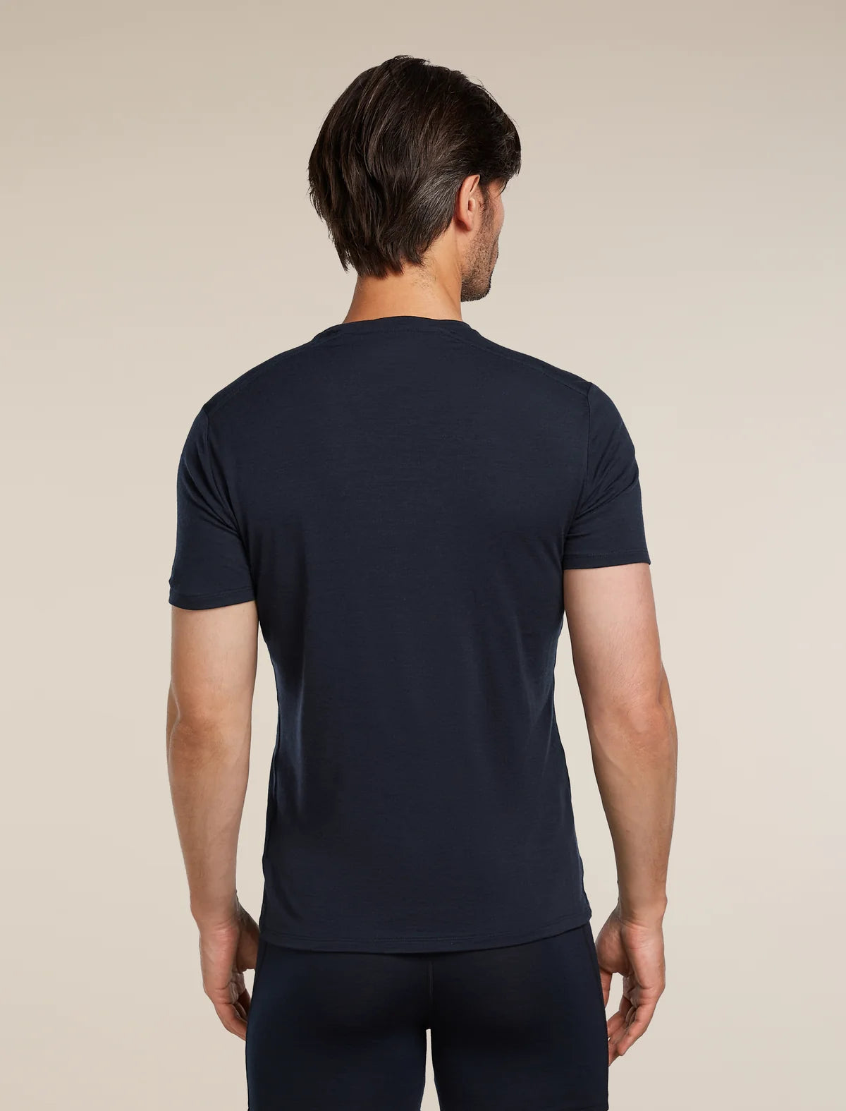A man viewed from the back wearing a dark navy blue short-sleeve crewneck t-shirt and matching dark pants, set against a plain beige studio background. - Icebreaker Merino Anatomica Short Sleeve Crew Neck Tee (Men's) - Midnight Tee