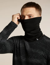 A man wearing a black neck gaiter balaclava, pulling it up over his nose and mouth while wearing a black speckled long-sleeve shirt, against a plain beige background. - Icebreaker Merino 260 Apex Chute (Unisex) - Black