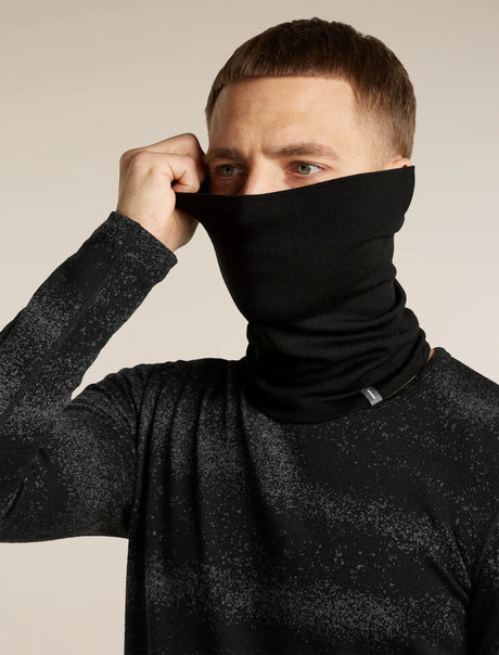 A man wearing a black neck gaiter balaclava, pulling it up over his nose and mouth while wearing a black speckled long-sleeve shirt, against a plain beige background. - Icebreaker Merino 260 Apex Chute (Unisex) - Black