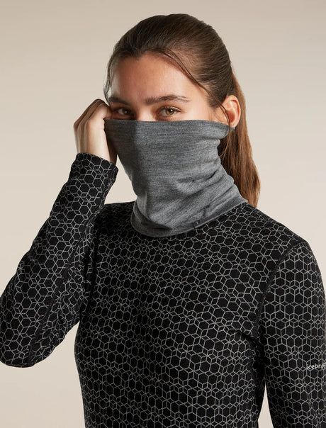 A woman wearing a dark gray heathered neck gaiter mask and a black long-sleeve athletic top with a white geometric honeycomb pattern, set against a solid beige background. - Icebreaker Merino 260 Apex Chute (Unisex) - Gritstone Heather