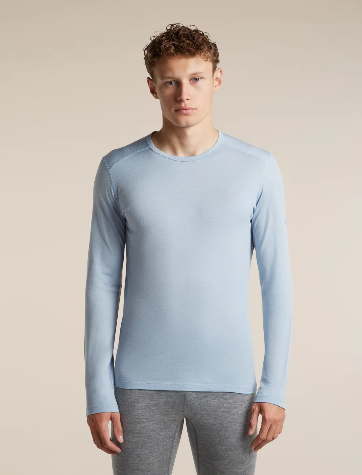 A man wearing a light blue long-sleeve crewneck athletic shirt and gray leggings, standing against a solid beige studio background. - Icebreaker Merino 200 Oasis Long Sleeve Crew Thermal Top (Men's) - Zinc Blue