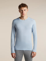 A man wearing a light blue long-sleeve crewneck athletic shirt and gray leggings, standing against a solid beige studio background. - Icebreaker Merino 200 Oasis Long Sleeve Crew Thermal Top (Men's) - Zinc Blue