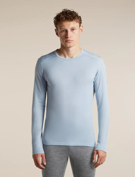 A man wearing a light blue long-sleeve crewneck athletic shirt and gray leggings, standing against a solid beige studio background. - Icebreaker Merino 200 Oasis Long Sleeve Crew Thermal Top (Men's) - Zinc Blue