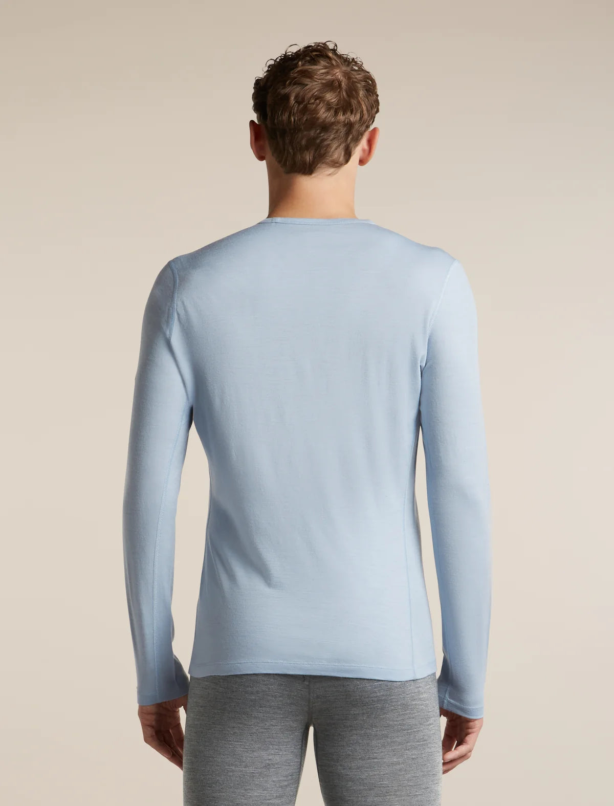 A man viewed from the back wearing a light blue long-sleeve crewneck shirt and gray heather pants, set against a plain beige studio background. - Icebreaker Merino 200 Oasis Long Sleeve Crew Thermal Top (Men's) - Zinc Blue