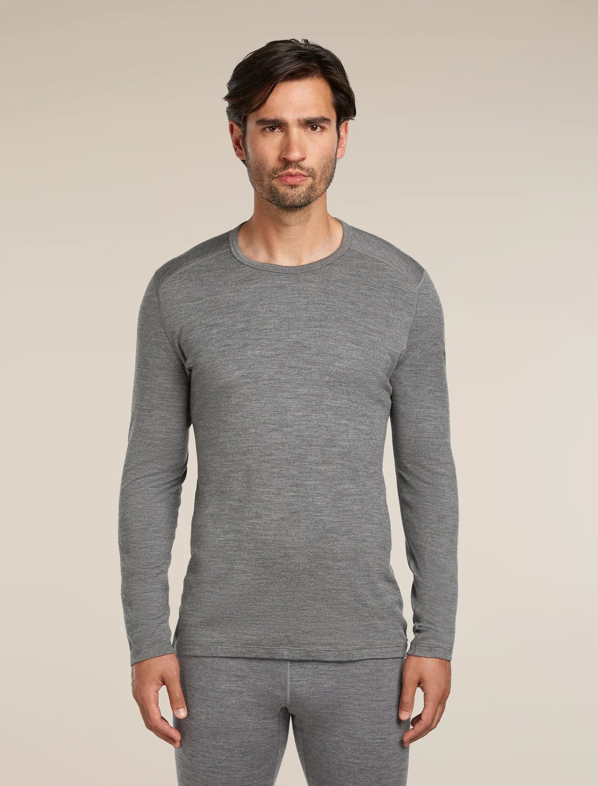 A man wearing a heather gray long-sleeve crewneck shirt and matching gray pants, standing against a plain beige studio background. - Icebreaker Merino 260 Oasis Long Sleeve Crew Thermal Top (Men's) - Gritstone Heather
