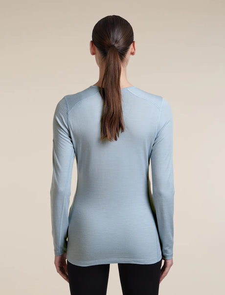 A woman viewed from the back wearing a light blue long-sleeve athletic top and black leggings against a plain beige studio background. - Icebreaker Merino 200 Oasis Long Sleeve Crew Thermal Top (Women's) - Zinc Blue