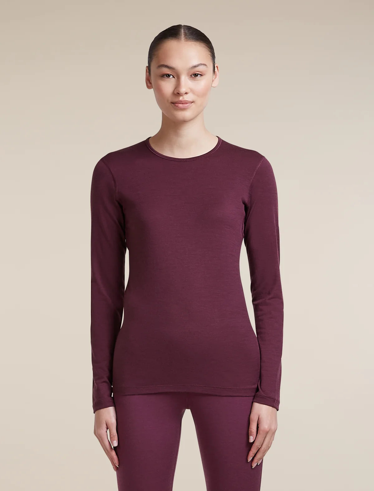 A woman modeling a long-sleeve crewneck top and matching leggings in a deep plum purple color, set against a solid beige studio background. - Icebreaker Merino 200 Oasis Long Sleeve Crew Thermal Top (Women's) - Dark Berry