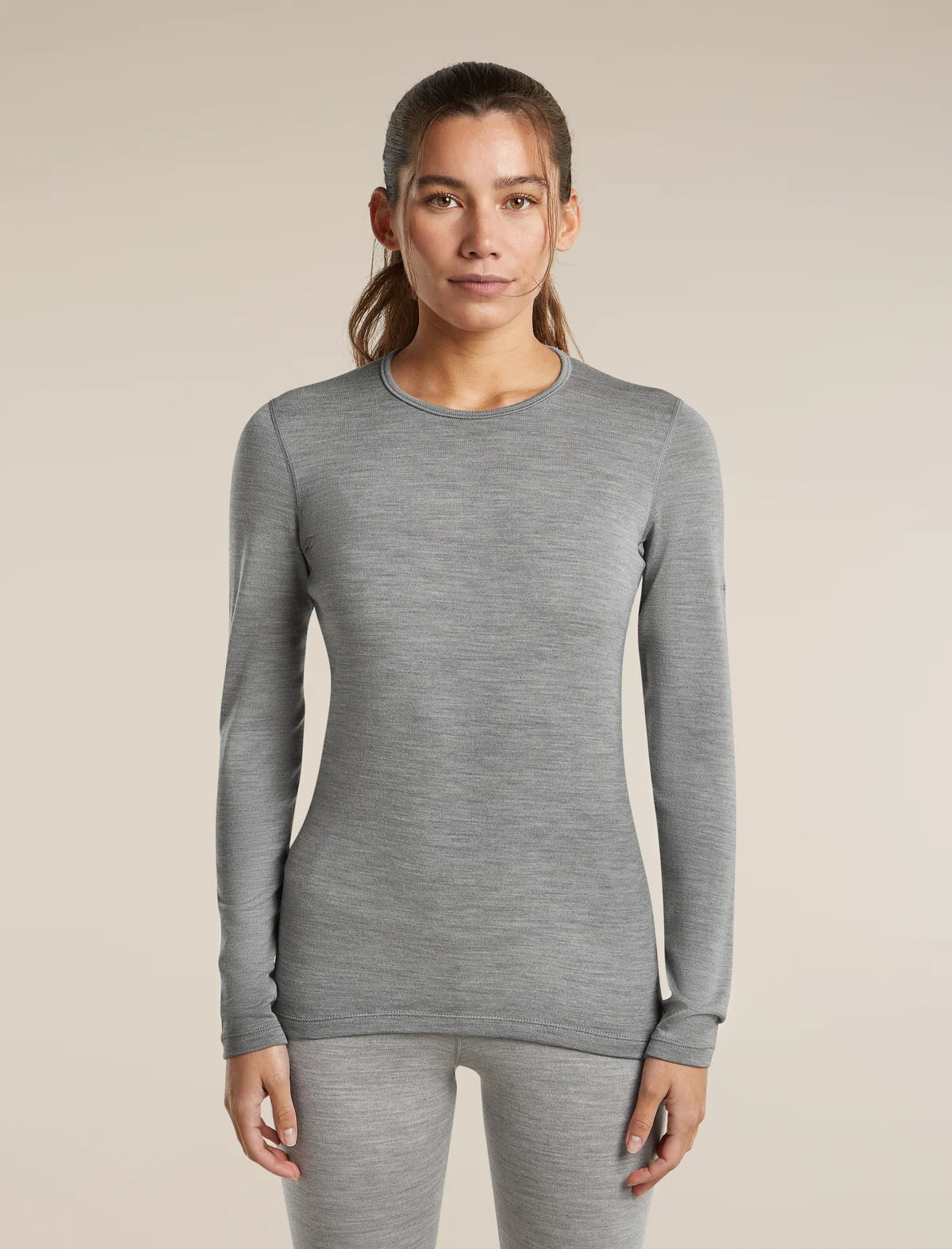 A woman modeling a fitted long-sleeve crewneck top in heather gray, paired with matching gray leggings, against a plain beige studio background. - Icebreaker Merino 260 Oasis Long Sleeve Crew Thermal Top (Women's) - Metro Heather