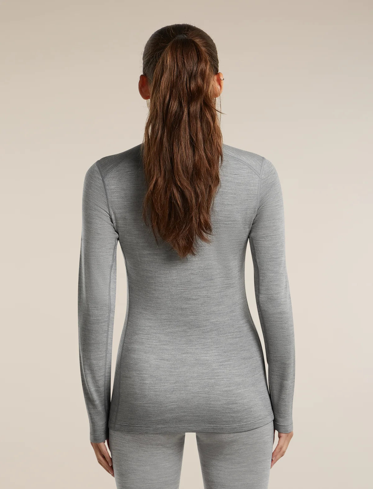 A woman viewed from the back wearing a fitted long-sleeve heather gray top and matching gray bottoms against a plain warm beige studio background. - Icebreaker Merino 260 Oasis Long Sleeve Crew Thermal Top (Women's) - Metro Heather
