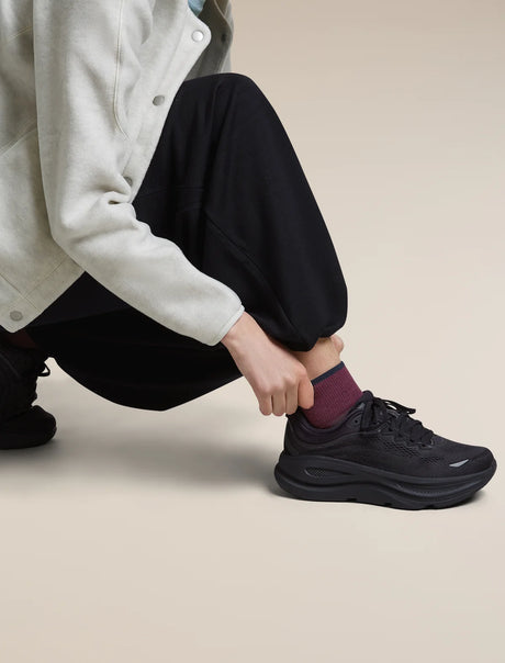 A person crouching and pulling up a black athletic sneaker with a chunky sole, wearing black pants, a light gray cardigan, and a burgundy sock, against a plain beige studio background. - Icebreaker Merino Multisport Light Mini Socks (Women's) - Dark Berry/Midnight Navy/Grey Plum