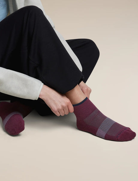 A person seated cross-legged wearing dark pants and low-cut socks in a burgundy and gray knit pattern, with one hand adjusting the ankle sock. - Icebreaker Merino Multisport Light Mini Socks (Women's) - Dark Berry/Midnight Navy/Grey Plum