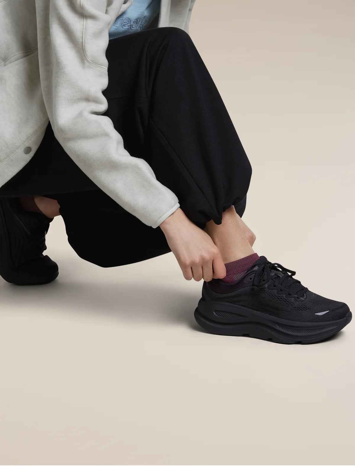A person crouching while putting on a black athletic sneaker with a chunky sole, paired with black jogger pants and a light gray hoodie, against a solid beige background. - Icebreaker Merino Multisport Light Micro Socks (Women's) - Dark Berry/Midnight Navy/Grey Plum