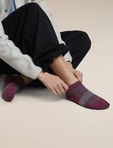 A person sitting on a light beige floor wearing dark pants and light gray sleeves, adjusting a low-cut knit sock on one foot. The sock is burgundy with a gray knit stripe across the instep. - Icebreaker Merino Multisport Light Micro Socks (Women's) - Dark Berry/Midnight Navy/Grey Plum