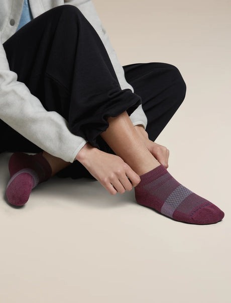 A person sitting on a light beige floor wearing dark pants and light gray sleeves, adjusting a low-cut knit sock on one foot. The sock is burgundy with a gray knit stripe across the instep. - Icebreaker Merino Multisport Light Micro Socks (Women's) - Dark Berry/Midnight Navy/Grey Plum