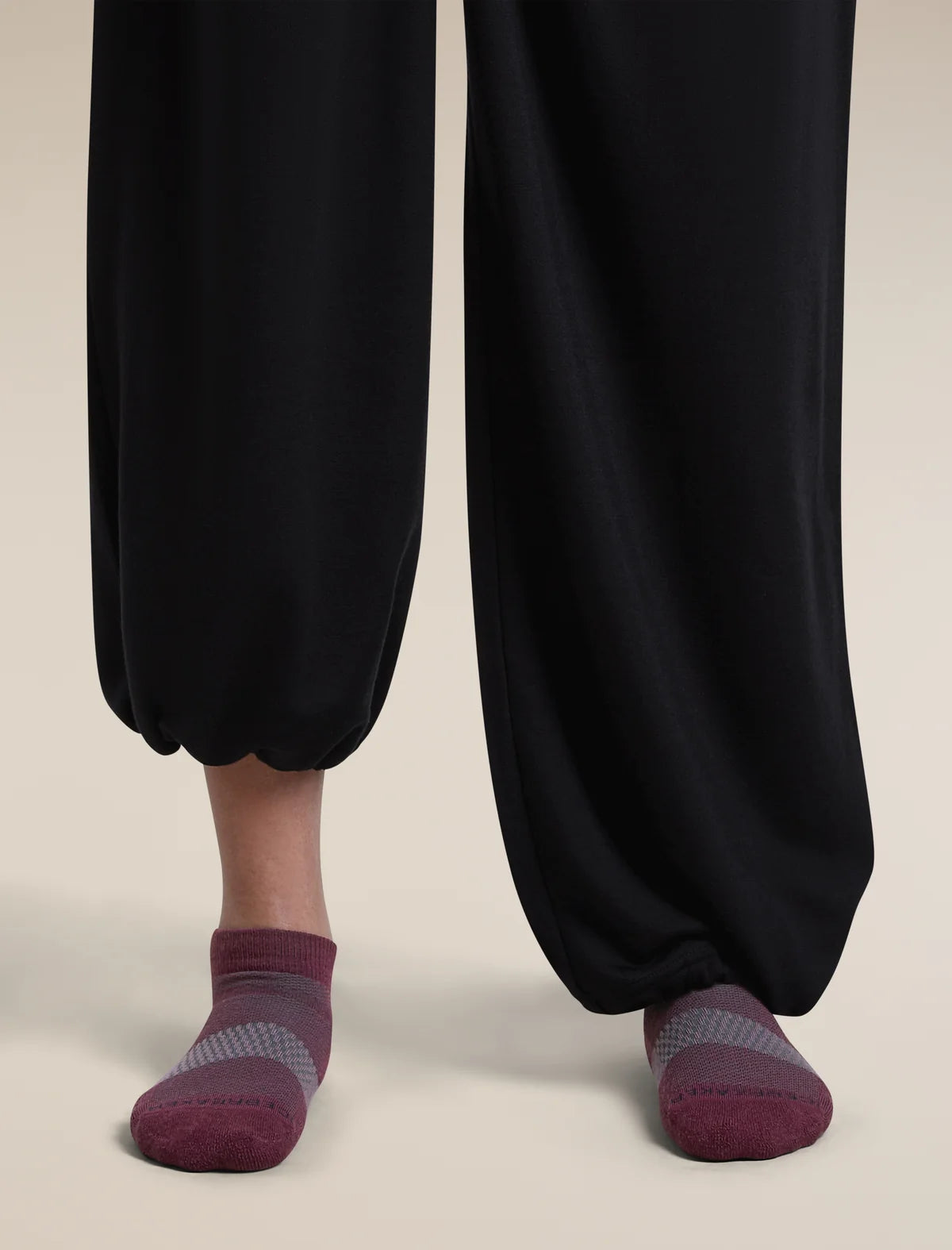 A close-up of a person's feet wearing dark charcoal black harem-style pants and burgundy low-cut socks with gray striped detailing, set against a solid light beige background. - Icebreaker Merino Multisport Light Micro Socks (Women's) - Dark Berry/Midnight Navy/Grey Plum