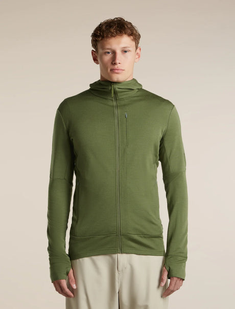 A man wearing an olive green full-zip hooded jacket with a vertical zip chest pocket, paired with light beige trousers, standing against a solid beige studio background. - Icebreaker Merino 260 Quantum Long Sleeve Zip Hoodie (Men's) - Sphagnum