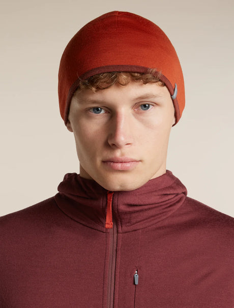A close-up portrait of a man wearing a rust-orange knit beanie and a matching dark burgundy zip-up fleece jacket against a plain beige studio background. - Icebreaker Merino 200 Pocket Hat (Unisex) - Agate/Port