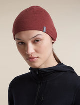 A woman wearing a dark red knit beanie with a small blue brand tag and a black zip-up hoodie, photographed against a solid beige background. - Icebreaker Merino 200 Pocket Hat (Unisex) - Agate/Port