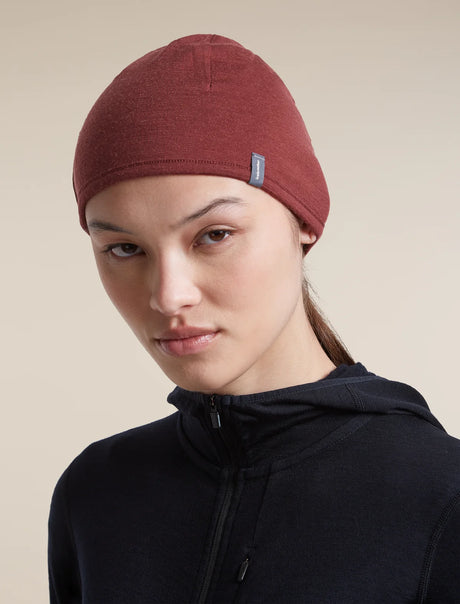 A woman wearing a dark red knit beanie with a small blue brand tag and a black zip-up hoodie, photographed against a solid beige background. - Icebreaker Merino 200 Pocket Hat (Unisex) - Agate/Port