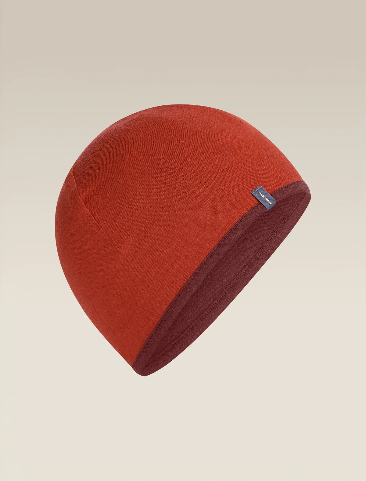 A knit beanie hat in a warm rust red color, shown floating against a solid light beige background. A small dark blue brand tag is visible on the side. - Icebreaker Merino 200 Pocket Hat (Unisex) - Agate/Port