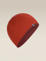 A knit beanie hat in a warm rust red color, shown floating against a solid light beige background. A small dark blue brand tag is visible on the side. - Icebreaker Merino 200 Pocket Hat (Unisex) - Agate/Port