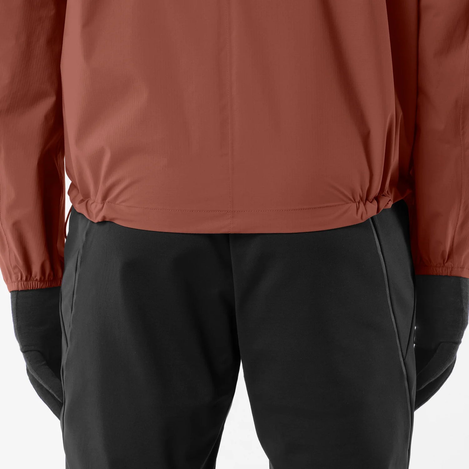 Close-up of a person wearing a rust-brown lightweight jacket and black pants, with black gloves visible against a clean white studio background. - Salomon Bonatti Waterproof Jacket (Men's) - Burnt Umber