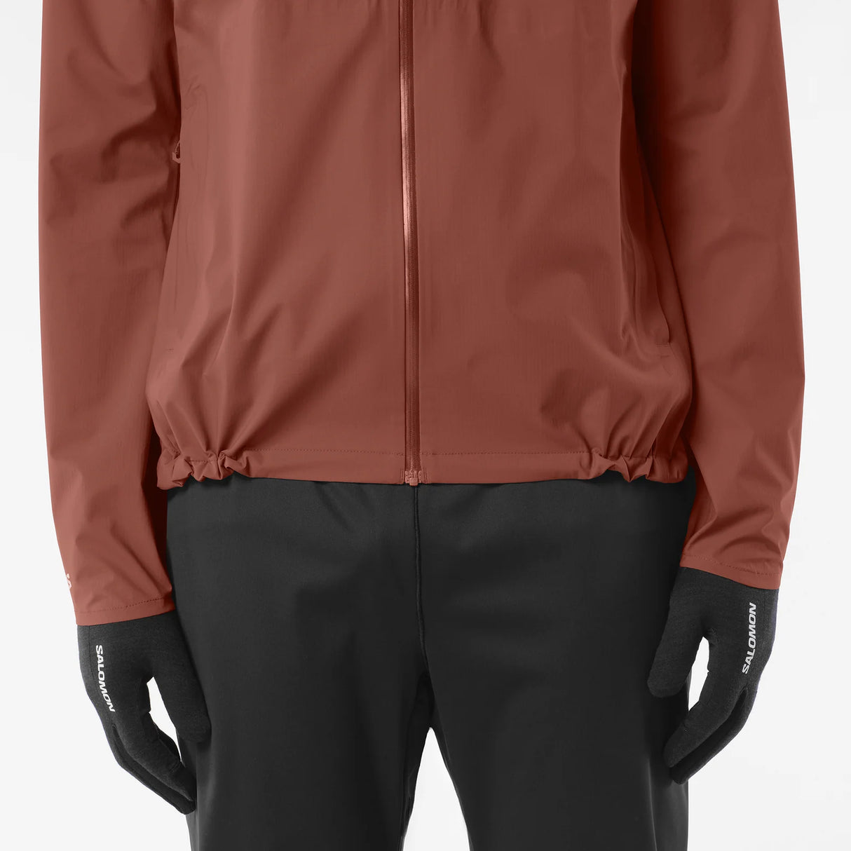 A close-up of a person wearing a rust-brown zip-up jacket, black pants, and black gloves with white 'SALOMON' branding on the cuffs, against a light gray studio background. - Salomon Bonatti Waterproof Jacket (Men's) - Burnt Umber