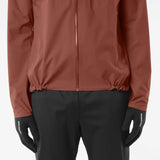 A close-up of a person wearing a rust-brown zip-up jacket, black pants, and black gloves with white 'SALOMON' branding on the cuffs, against a light gray studio background. - Salomon Bonatti Waterproof Jacket (Men's) - Burnt Umber