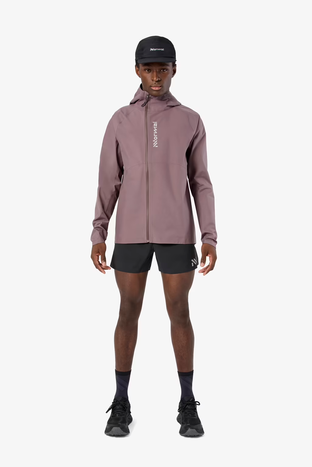 A male model wearing a dusty rose zip-up hooded jacket with a vertical white 'ROCKNROLLER' logo on the chest, black running shorts, a black cap, and black running shoes, standing against a light gray background. - NNormal Trail Rain Jacket 02 (Men's) - Purple