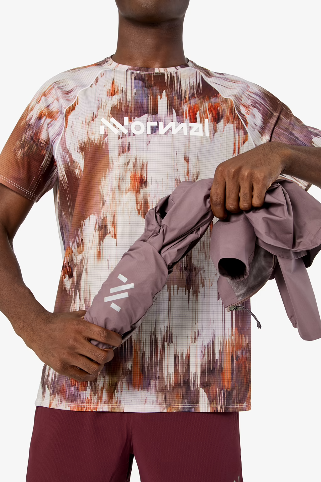 A close-up of a model wearing a short-sleeve athletic t-shirt with an abstract paint-splatter pattern in white, orange, and brown, holding a dusty purple jacket. The brand name 'Noguryal' is printed across the chest. - NNormal Trail Rain Jacket 02 (Men's) - Purple
