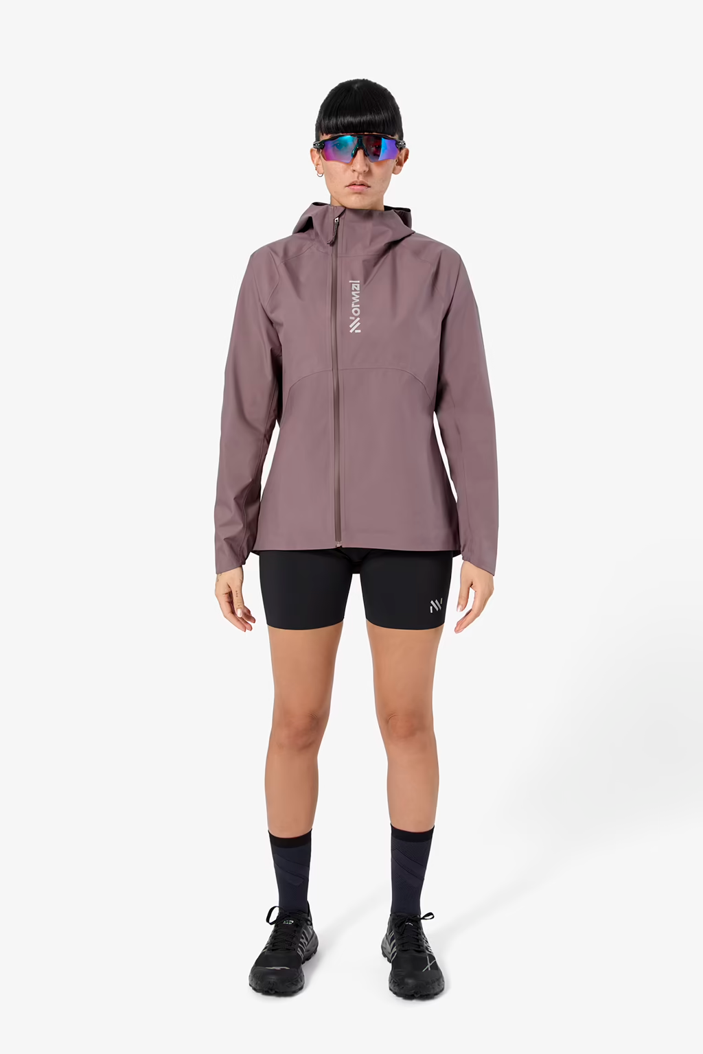 A model stands against a light gray studio background wearing a muted mauve-purple hooded athletic jacket with a vertical white 'NORDIC' logo on the chest, black running shorts, black socks, and black trail running shoes. - NNormal Trail Rain Jacket 02 (Women's) - Purple