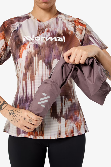 A woman wearing a short-sleeve athletic jersey with an abstract multicolor print in purple, brown, orange, and white, holding a matching dusty purple sleeve or sleeve protector in front of her torso. The jersey has the brand name 'NORIMAL' printed across the chest. - NNormal Trail Rain Jacket 02 (Women's) - Purple