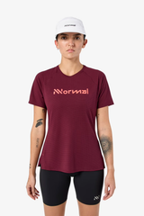 A woman modeling a maroon athletic t-shirt with a pink 'Normal' logo on the chest, paired with black bike shorts and a white cap, against a light gray background. - NNormal Race T-Shirt 02 (Women's) - Burgundy