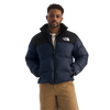 A man wearing a navy blue and black puffer jacket with a white embroidered The North Face logo on the chest, paired with tan pants, against a solid white background. - The North Face 1996 Retro Nuptse Jacket (Men's) - Summit Navy/TNF Black