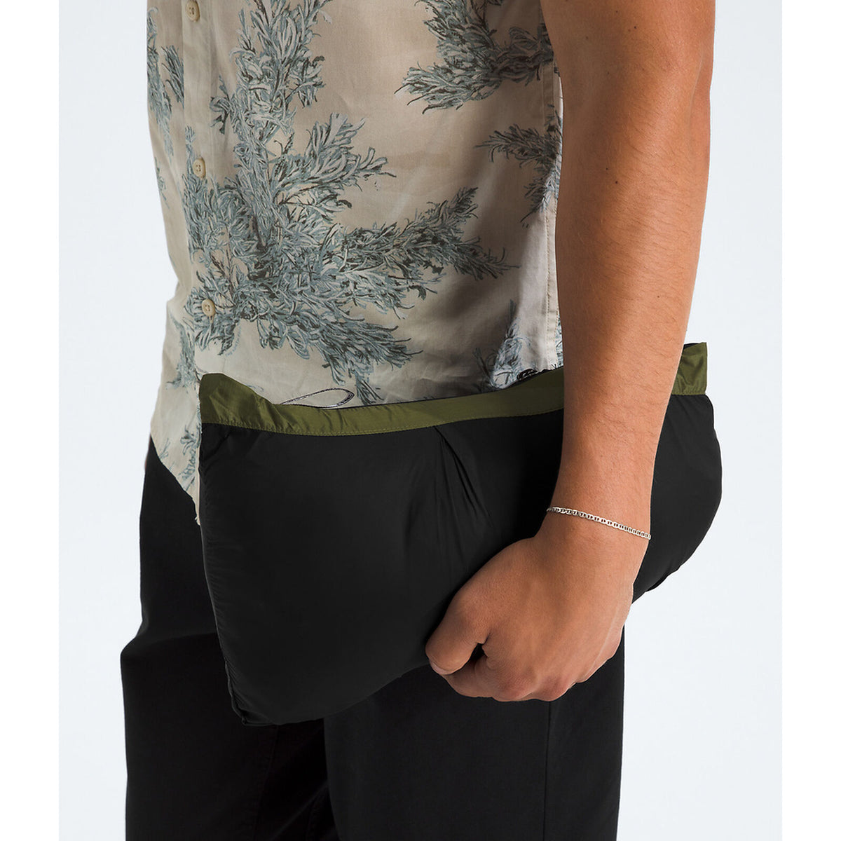 Close-up of a person wearing a black pouch-style garment with an olive green waistband, paired with a beige patterned shirt and black pants, against a light gray background. - The North Face 1996 Retro Nuptse Jacket (Men's) - Woodland Green/TNF Black