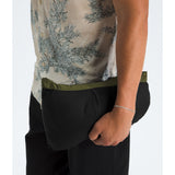 Close-up of a person wearing a black pouch-style garment with an olive green waistband, paired with a beige patterned shirt and black pants, against a light gray background. - The North Face 1996 Retro Nuptse Jacket (Men's) - Woodland Green/TNF Black