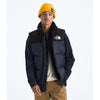 A man wearing a navy and black puffer vest by The North Face, a yellow beanie, a white t-shirt, and tan pants, standing against a light gray studio background. - The North Face 1996 Retro Nuptse Vest (Men's) - Summit Navy/TNF Black-Recycled Down