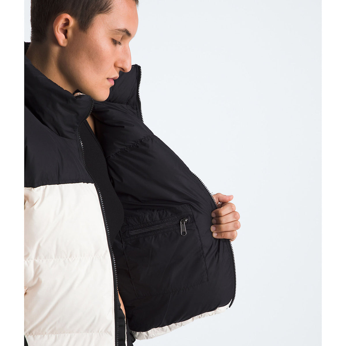 A close-up side view of a person wearing a black and white puffer jacket, opening the front to show a zippered interior pocket. - The North Face 1996 Retro Nuptse Jacket (Womens) - White Dune/TNF Black-Recycled Down