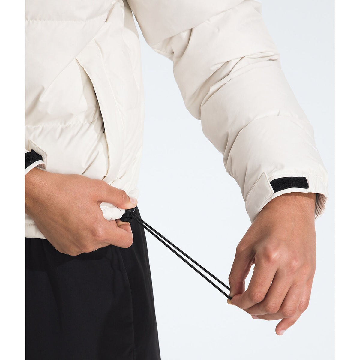 Person adjusting a black drawstring on a white jacket against a light gray background - The North Face 1996 Retro Nuptse Jacket (Womens) - White Dune/TNF Black-Recycled Down
