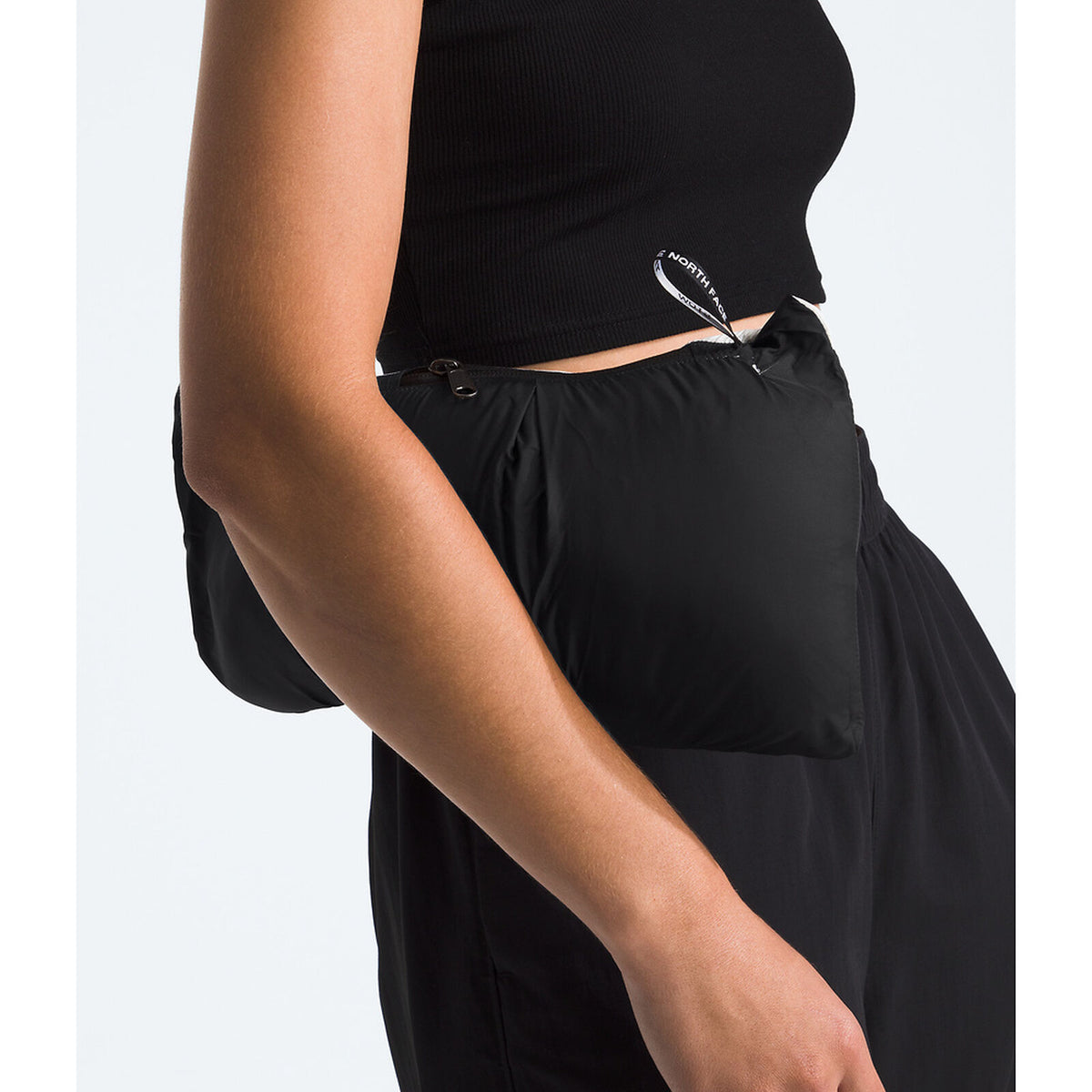 A close-up of a person wearing a black ribbed crop top and black wide-leg pants, with a small black pouch or pocket attached at the side waist with a silver metal clip. - The North Face 1996 Retro Nuptse Jacket (Womens) - White Dune/TNF Black-Recycled Down