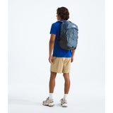 A man wearing a blue t-shirt, beige shorts, and running shoes is shown from behind, carrying a blue and black The North Face backpack on his back against a white studio background. - The North Face Borealis Backpack - Winslor Blue/Anthracite Grey/Utility Brown