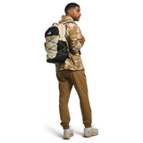 A man wearing a beige and brown camouflage jacket, brown pants, and white sneakers, shown from the back wearing a black and beige backpack with white cording. The photo is shot against a solid white background. - The North Face Borealis Backpack - Desert Stone/Stone Slab/Ember Soil